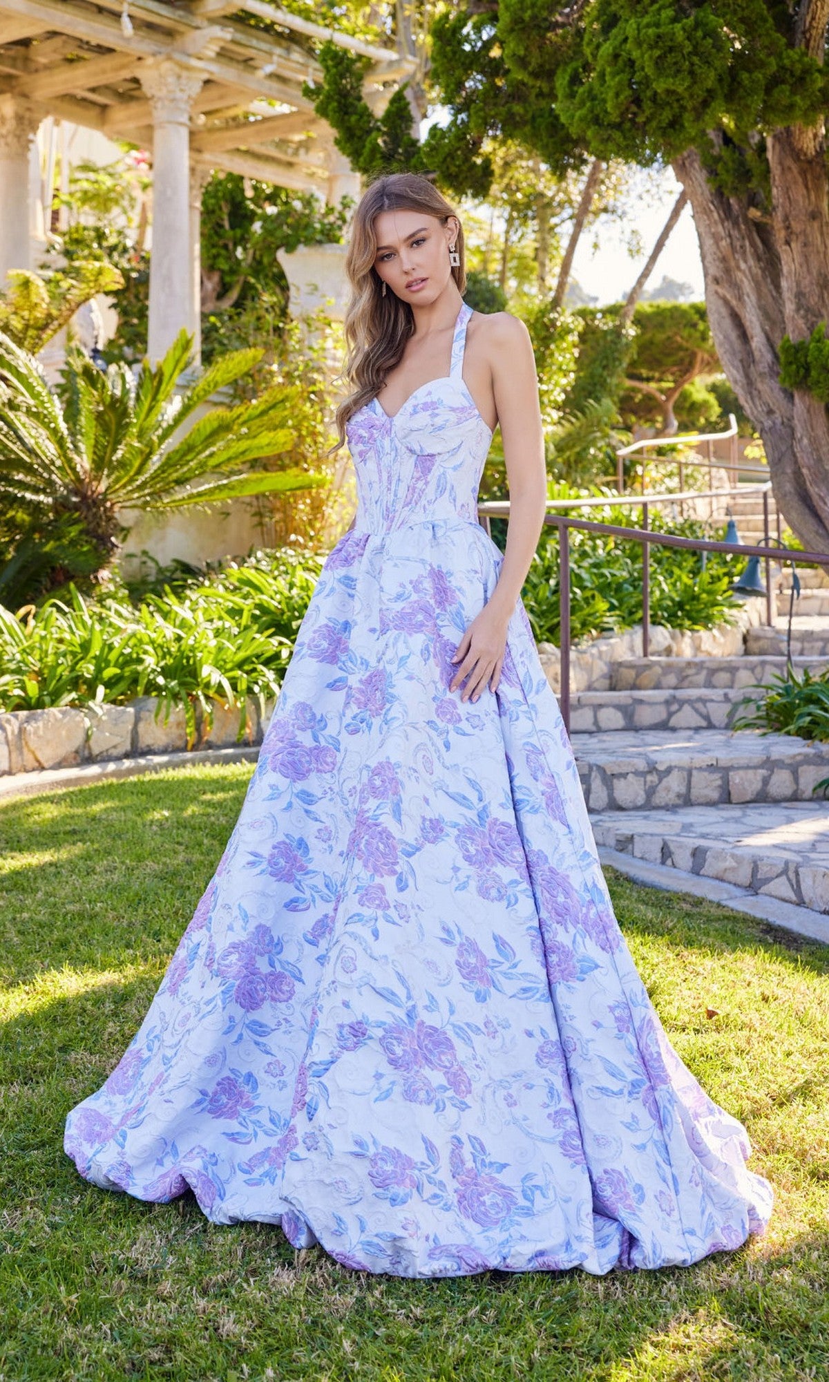 A woman stands outdoors on grass wearing the Juliet JT2742A Long Prom Dress in Lavender Amethyst, featuring a floral-textured design, corset bodice, and halter neckline; trees and a stone path are in the background.